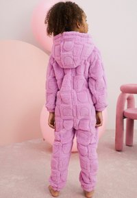 Combinaison en velours violet avec capuche, présentant un motif carré texturé. Manches et jambes ajustées, portée pieds nus, sur un fond rose doux.