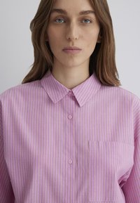 Light pink button-up shirt with vertical white and purple stripes, collared neckline, and single chest pocket. Smooth fabric texture.