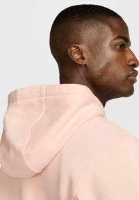 Felpa rosa chiaro con cappuccio regolabile, texture morbida e dettagli di cucitura visibili. La vista laterale mostra un orecchino discreto.