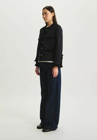 Veste noire texturée avec col rond, poches avant et fermeture à boutons. Associée à un jean bleu taille haute et des chaussures noires.