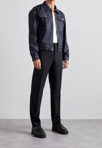 Hombre vestido con una chaqueta de mezclilla azul oscura, camiseta blanca, pantalones negros a medida y mocasines de cuero negros, de pie sobre un suelo gris.
