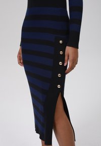 Kläddress i stickad kvalitet med marint och svart horisontella ränder, slits på sidan och guldknappdetaljer på ena sidan. Smejig textur, åtsittande design.