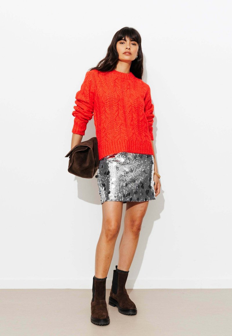 Pull rouge en maille torsadée, mini-jupe argentée à sequins, bottines en daim marron et pochette marron. Surfaces lisses et texturées.