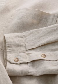 Beige linnen shirtstof met zichtbare textuur. Voorzien van een met knoopsluiting verwisselbare manchette met een lichtbruin knoopje en een omgevouwen zoom.