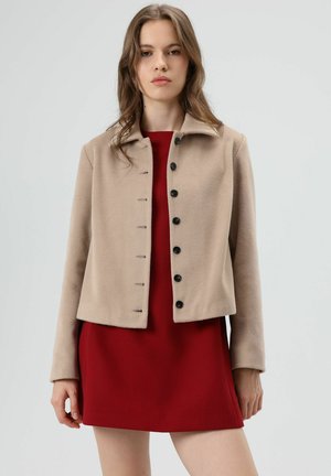 Beige, cropped, gestructureerd jasje met een kraag, voorzien van zwarte knopen aan de voorkant. Draagt over een rode jurk en heeft een eenvoudig, strak ontwerp.