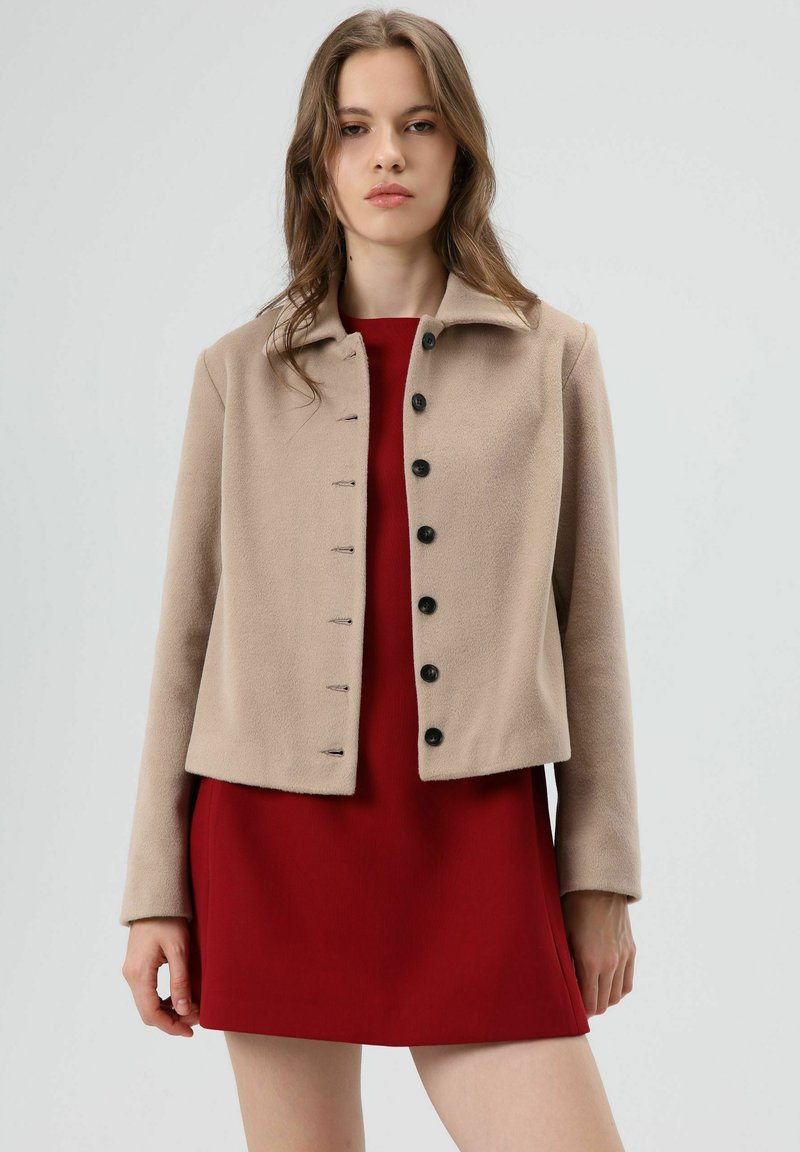 Beige, cropped, gestructureerd jasje met een kraag, voorzien van zwarte knopen aan de voorkant. Draagt over een rode jurk en heeft een eenvoudig, strak ontwerp.