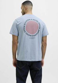 Šviesiai mėlyna medvilninė marškinė su apvaliu raštuotuoju grafiką raudonos ir juodos spalvos, aplink dizainą yra užrašas "Royal Denim Division".