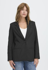 Črn blazer z enojnim zapiranjem na gumb, z navadnimi reverji, dvema sprednjima žepoma in prilegajočim se krojem. Obut je čez belo majico in modre kavbojke.