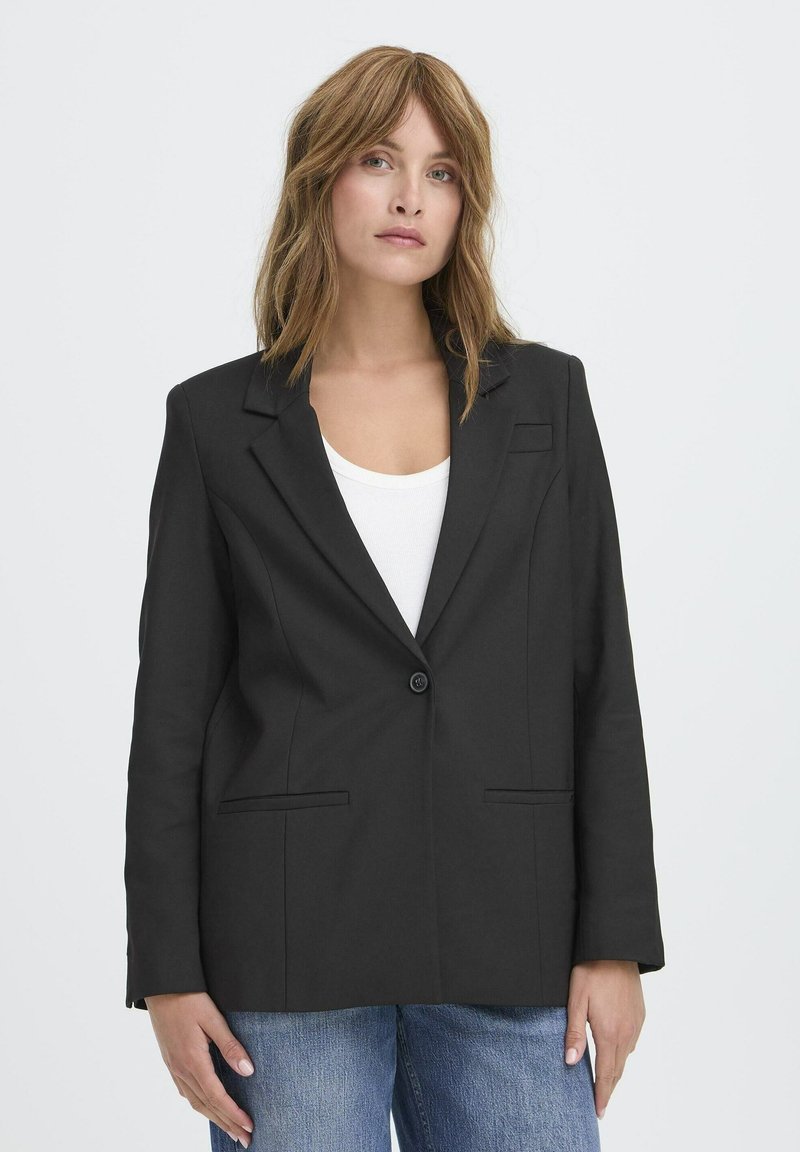 Črn blazer z enojnim zapiranjem na gumb, z navadnimi reverji, dvema sprednjima žepoma in prilegajočim se krojem. Obut je čez belo majico in modre kavbojke.