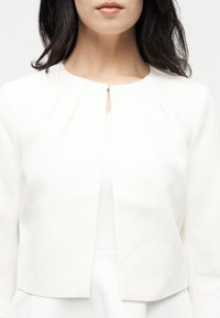 Femme portant une veste courte blanche à manches longues avec des plis près de l'encolure et une petite fermeture centrale à agrafe.