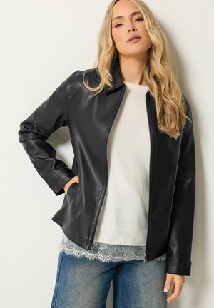 Donna che indossa una giacca di pelle nera sopra un maglione bianco lavorato a maglia con bordo in pizzo, abbinata a jeans blu, in piedi davanti a uno sfondo chiaro e semplice.