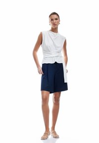 Witte mouwloze top met een gedrapeerd ontwerp en een zijstrik, gecombineerd met marineblauwe shorts. Vlotte stoftextuur, minimalistische stijl, geen patronen.