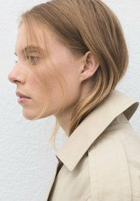 Profilvy av en person med rakt, ljusbrunt hår som bär en beige trenchcoat. Kappan har en bred krage och rena linjer.