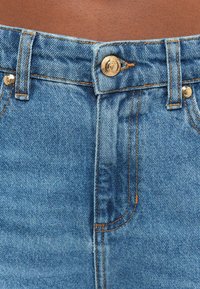 Jean en denim bleu avec une taille haute, fermeture à boutons dorés et coutures contrastantes orange, capturant la texture du tissu.