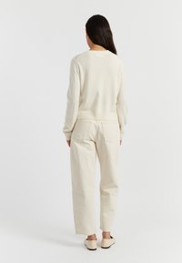 Maglione di colore chiaro a maglia con scollo rotondo, abbinato a pantaloni dritti color bianco sporco. Entrambi gli articoli presentano una texture morbida e un design essenziale.