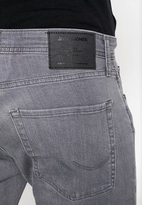 Grå jeans med en texturerad finish, kommer med en svart etikett vid midjan, funktionella bakfickor och sömdetaljer längs sömmarna.
