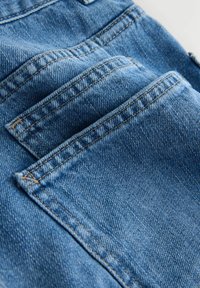 Niebieska tkanina dżinsowa z widocznymi przeszyciami i wieloma kieszeniami, lekko pognieciona, ukazująca fakturę i głębię koloru.