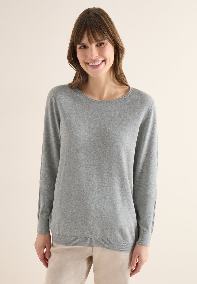 Femme souriante aux cheveux bruns portant un pull à manches longues gris clair et un pantalon beige, debout devant un fond clair uni.