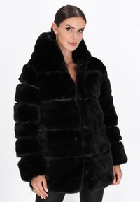 Cappotto in pelliccia sintetica nera con cappuccio, caratterizzato da un design squadrato, sezioni trapuntate e una texture liscia. Si chiude frontalmente senza hardware visibile.