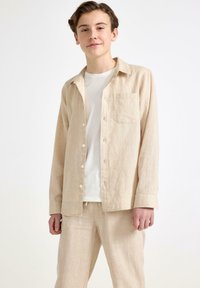 Camicia di lino beige chiaro con maniche lunghe, chiusura a bottoni e taschino sul petto, abbinata a pantaloni in lino coordinati. T-shirt bianca semplice sotto.