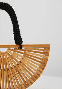 Sac à main en bois semi-circulaire à lattes avec une poignée épaisse noire enveloppée, présenté sur un fond gris clair uni.