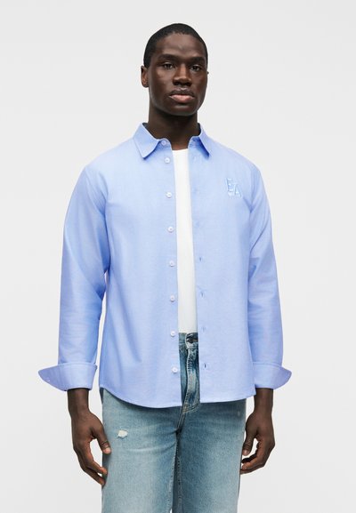 Chemise bleu clair à manches longues avec boutons sur le devant, col et logo brodé. Portée sur un t-shirt blanc et associée à un jean clair délavé.