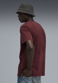 Marsala farvet bomulds T-shirt med korte ærmer, afslappet pasform, bæres med denim shorts. Grå bucket hat supplerer outfittet. Enkel design, ingen mønstre.