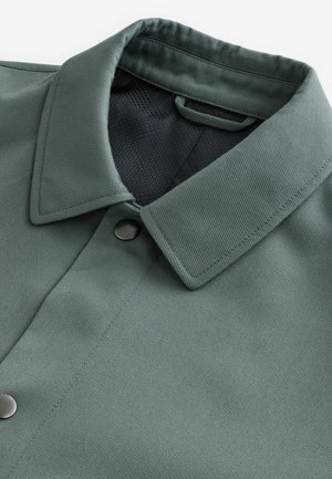 Camicia con colletto in tessuto verde tenue con una finitura texturizzata. Presenta un interno in rete nera e una chiusura a pulsante sul collo.