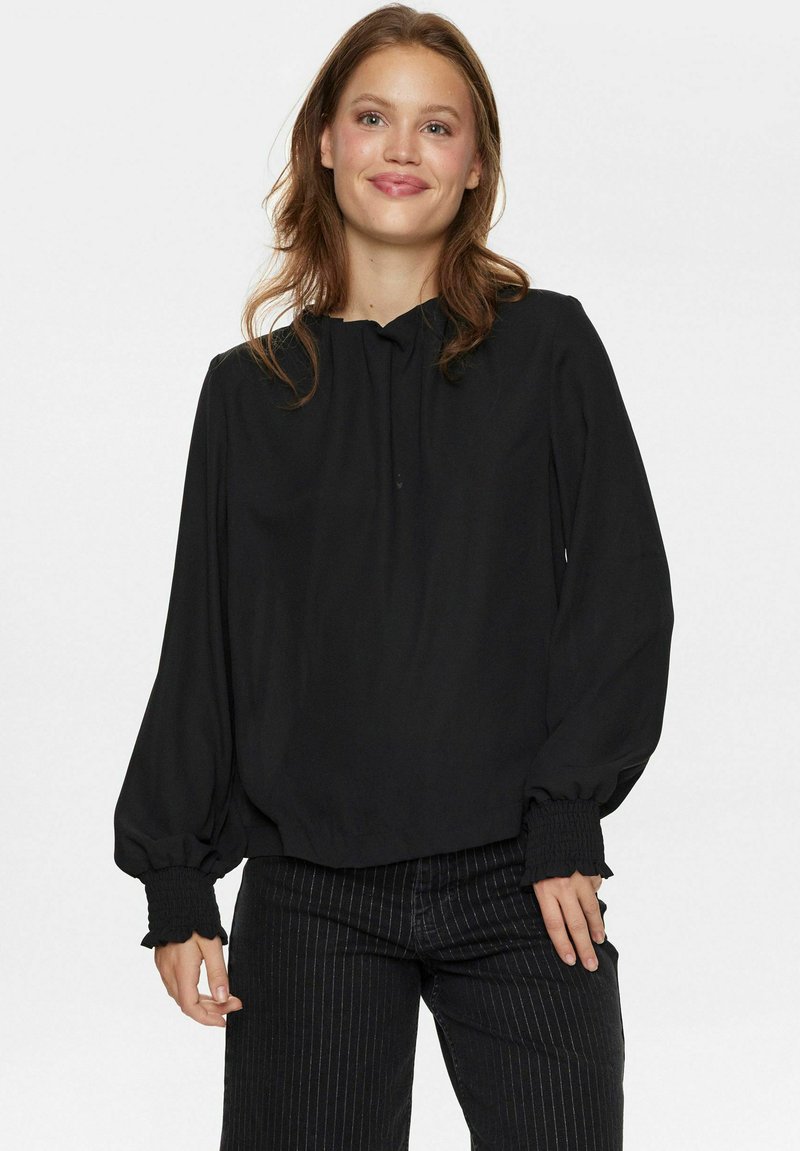 Blusa nera con maniche lunghe a sbuffo, scollatura rotonda e polsini elastici. Tessuto liscio con un leggero lucido. Abbinata a pantaloni neri a righe.