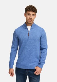 Lyseblå strikket sweater med ståkrave, lynlås foran og ribbede manchetter. Blød tekstur, tætsiddende design, parret med mørke jeans.