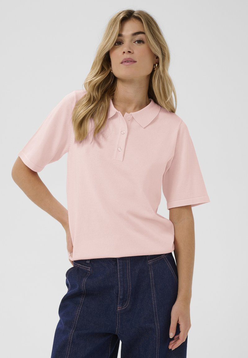 Polo rose clair avec une texture douce, manches courtes, col et patte de boutonnage à trois boutons. Porté avec un pantalon en denim foncé avec des coutures visibles.