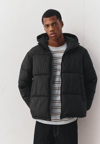 Nuori mies, jolla on musta toppatakki valkoisen ja mustaraidallisen paidan päällä, seisomassa yksinkertaisen vaalean seinän edessä.