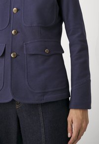 Chaqueta azul marino en tejido texturizado con cierres de botón y dos bolsillos frontales. Botones dorados destacados y diseño ajustado.