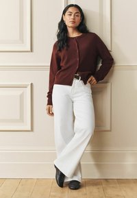 Tumši sarkans kardigans ar pogām priekšpusē, kombinēts ar baltām plaša piegriezuma džinsu biksēm un melniem apaviem. Gluda tekstūra, minimālistisks dizains pret neitrālu sienu.