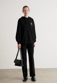 Svart oversized hettegenser med logo foran, kombinert med svarte rette bukser og skinnende svarte sko, utfylt med en liten svart håndveske.