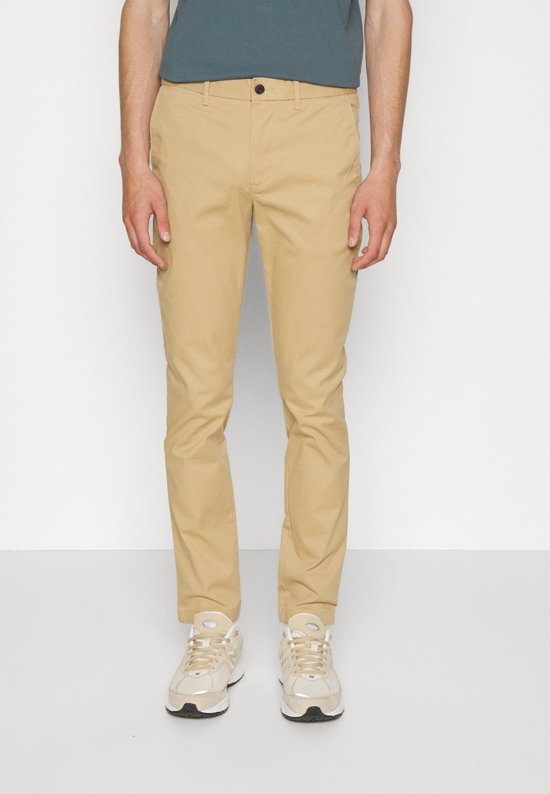 Khaki nohavice so Slim fit, vyrobené z bavlneného zloženia. Obsahujú predné vrecká, zapínanie na gombík a hladkú textúru. Skombinované s béžovými teniskami.