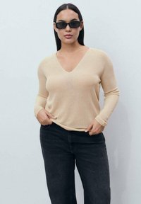 Beige V-hals trui met lange mouwen, gemaakt van textuur gebreid materiaal, gecombineerd met zwarte jeans. Model staat tegen een neutrale achtergrond.