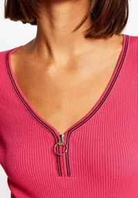 Top rose côtelé avec un décolleté en V et une fermeture éclair frontale dotée d'une tirette circulaire. Des surpiqûres noires soulignent le décolleté et la zone de la fermeture éclair.