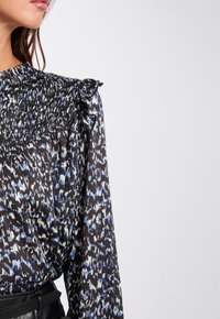 Schwarze Bluse mit blau-weißer abstrakter Musterung, mit gerafftem Ausschnitt und Rüschendetails an den Schultern. Der Stoff wirkt leicht und hat eine fließende Textur.