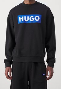 Felpa nera con colletto rotondo, caratterizzata da un grande logo "HUGO" blu e bianco sul davanti. Tessuto morbido, vestibilità ampia e polsini a coste.