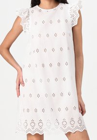 Witte linnen jurk met geborduurde bloemenuitsparingen, geruite mouwen en een afgezakte zoom. Lichtgewicht, ademende stof. Minimalistisch ontwerp.