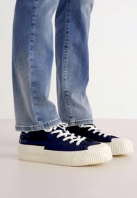 Navy canvas sneakers with white rubber soles, featuring prominent laces and textured toe caps, paired with light blue denim pants.