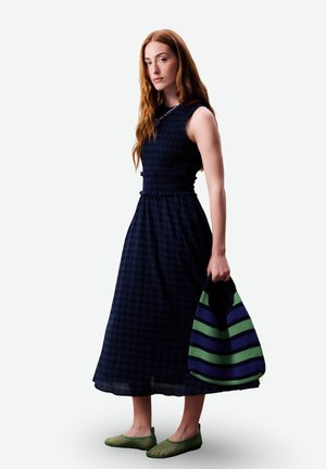 Frau mit langen roten Haaren, die ein ärmelloses, dunkelblau kariertes Midikleid trägt, eine grün-blau gestreifte Tasche hält und grüne Schuhe trägt.