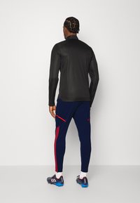 Černé dlouhé rukávy sportovní tričko, které je kombinováno s námořnickými sportovními kalhotami s červenými pruhy a akcentovanými kapsami, doplněné sportovními botami.