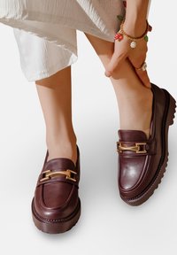 Mocassins en cuir bourgogne avec un bout rond, accents en métal doré et une semelle en caoutchouc texturée. Portés avec un tissu blanc fluide.