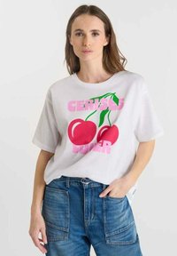 Femme portant un t-shirt blanc avec de grandes cerises rouges et le texte rose "Amoureuse des Cerises", accompagné de pantalons en denim bleu, se tenant devant un fond uni.