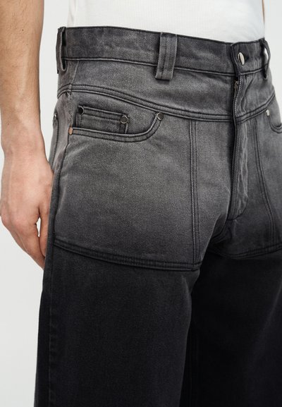 Άνδρας που φοράει τζιν ψηλόμεσο με διαβάθμιση από μαύρο σε γκρι, με ορατές ραφές και μεταλλικά καρφιά, με το χέρι ακουμπισμένο δίπλα του.