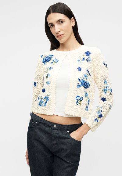 Γυναίκα που φοράει μια κρεμ πλεκτή ζακέτα με μπλε κεντήματα λουλουδιών πάνω από λευκό τοπ και σκούρα τζιν, στέκεται με το ένα χέρι στην τσέπη.