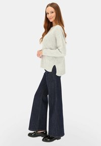 Kobieta w jasnoszarym swetrze z dzianiny, ciemnych szerokich dżinsach i czarnych lakierowanych mokasynach, stojąca w profilu z lekkim uśmiechem.