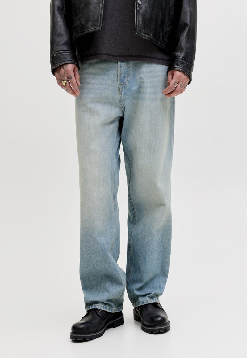 Personne portant un jean ample bleu clair, une veste en cuir noire, une chemise noire, des bottes noires et des bagues aux doigts sur fond blanc.